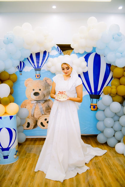 A woman in a traditional white outfit with a headpiece, standing in a room with balloons and a teddy bear, holding a cake, with a backdrop featuring a blue bear and the text 'Me and Mommy'.