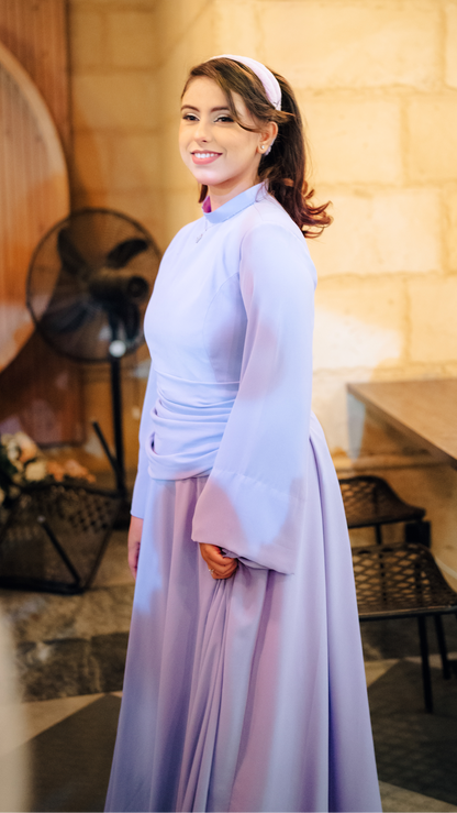 Woman in a light purple traditional outfit standing in a casual setting with a wooden wall and table.