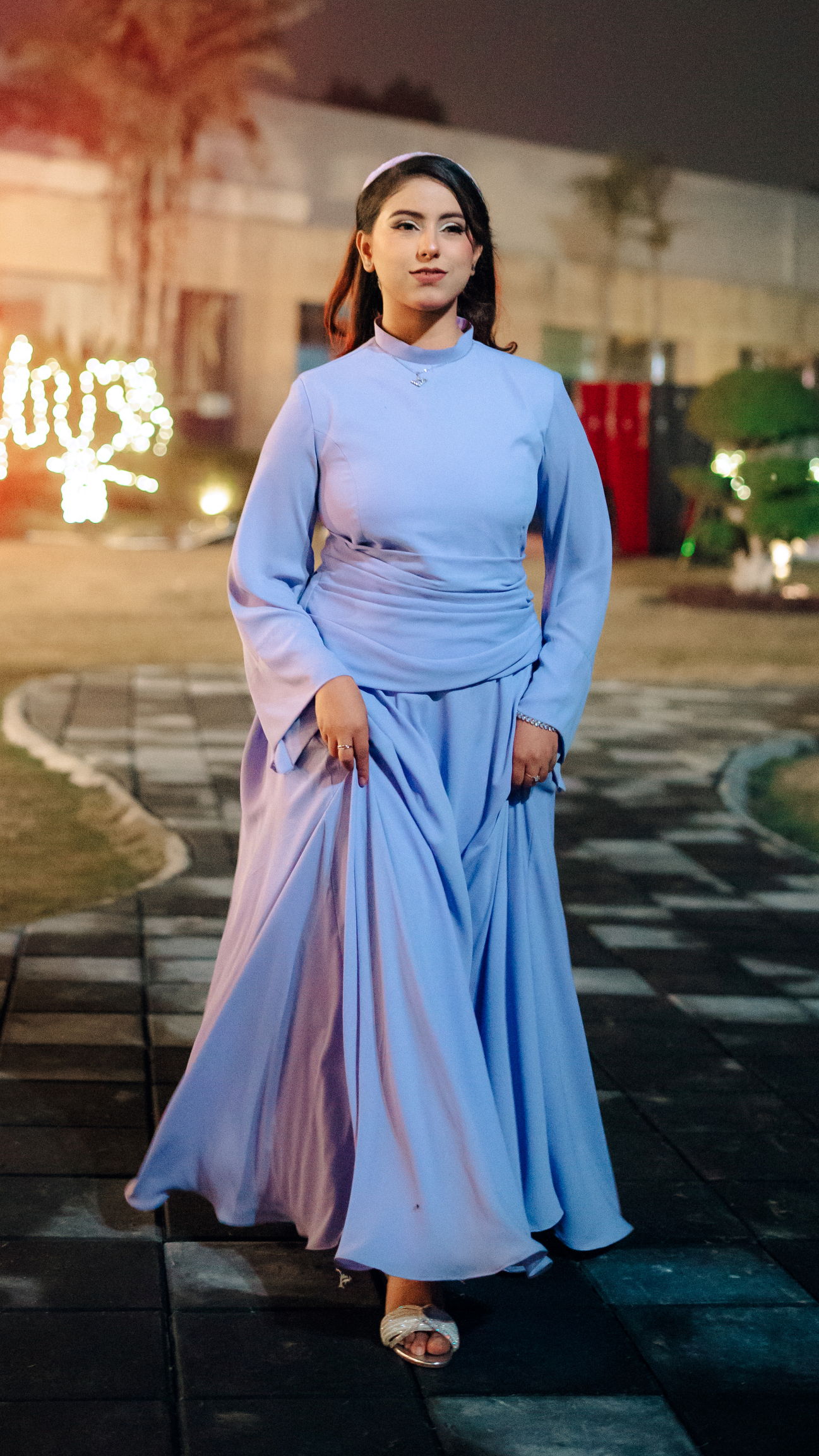 Woman in a light blue dress standing on a pathway at night with decorative lights in the background.