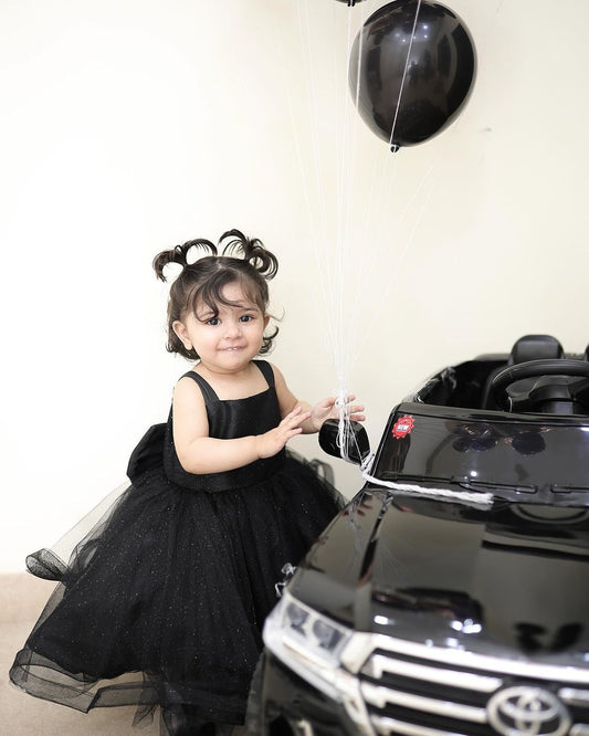 A child in a black dress with black hair, smiling and playing with a balloon near a black car.