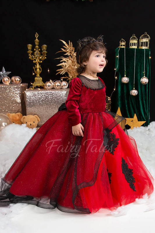 A child in a red velvet dress with black accents and decorative elements, standing in a setting with Christmas-themed decorations.