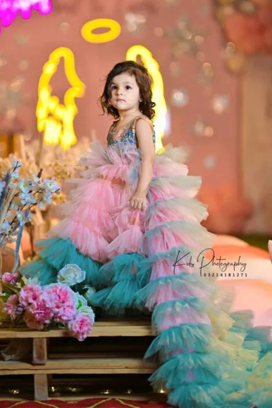 A young girl in a pink and blue floral dress with a delicate lace pattern, standing on a platform with decorative flowers and a backdrop featuring fairy lights.