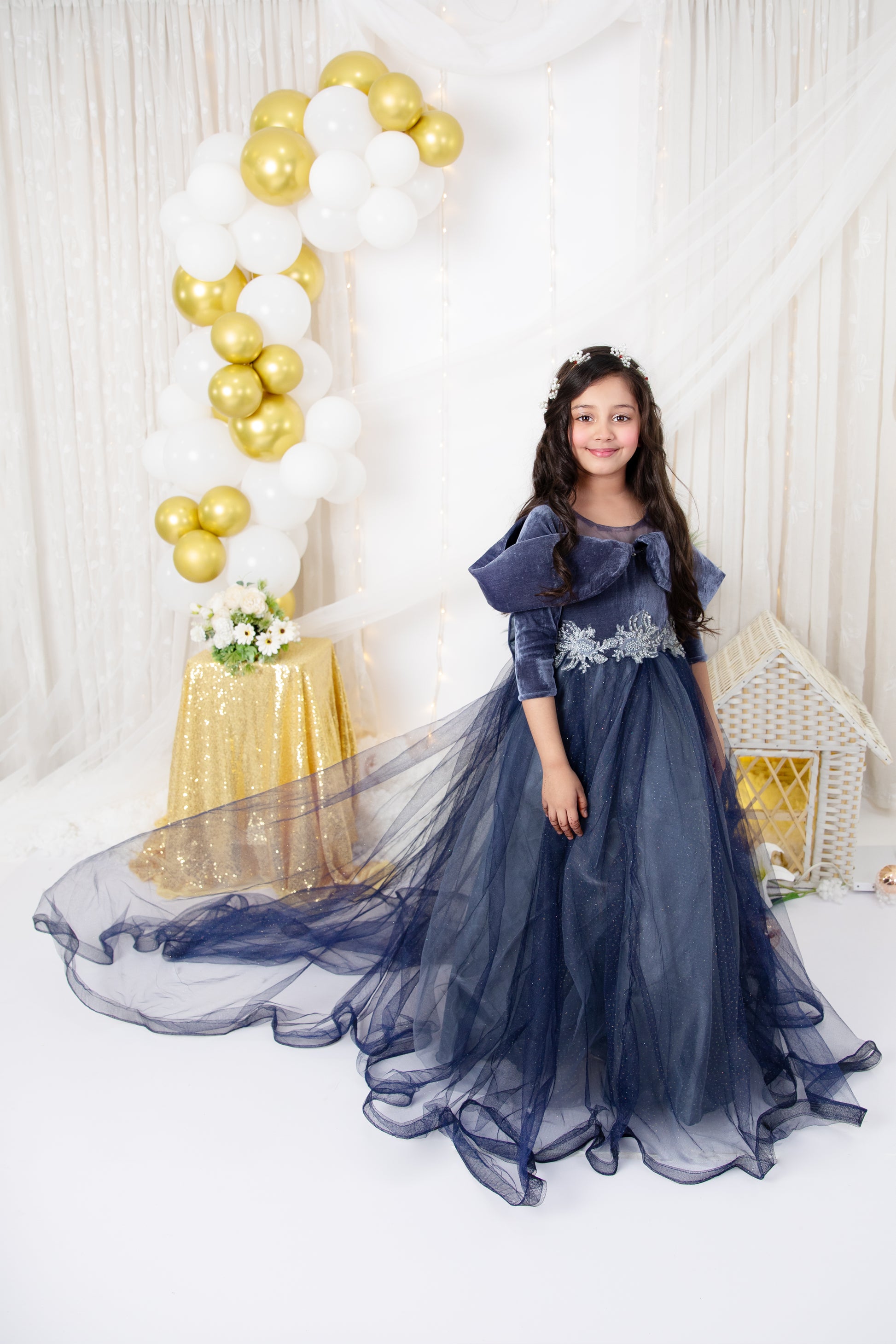 A young girl in a formal navy blue dress with a long train, featuring floral patterns and half-length sleeves, standing in front of a backdrop with golden balloons.